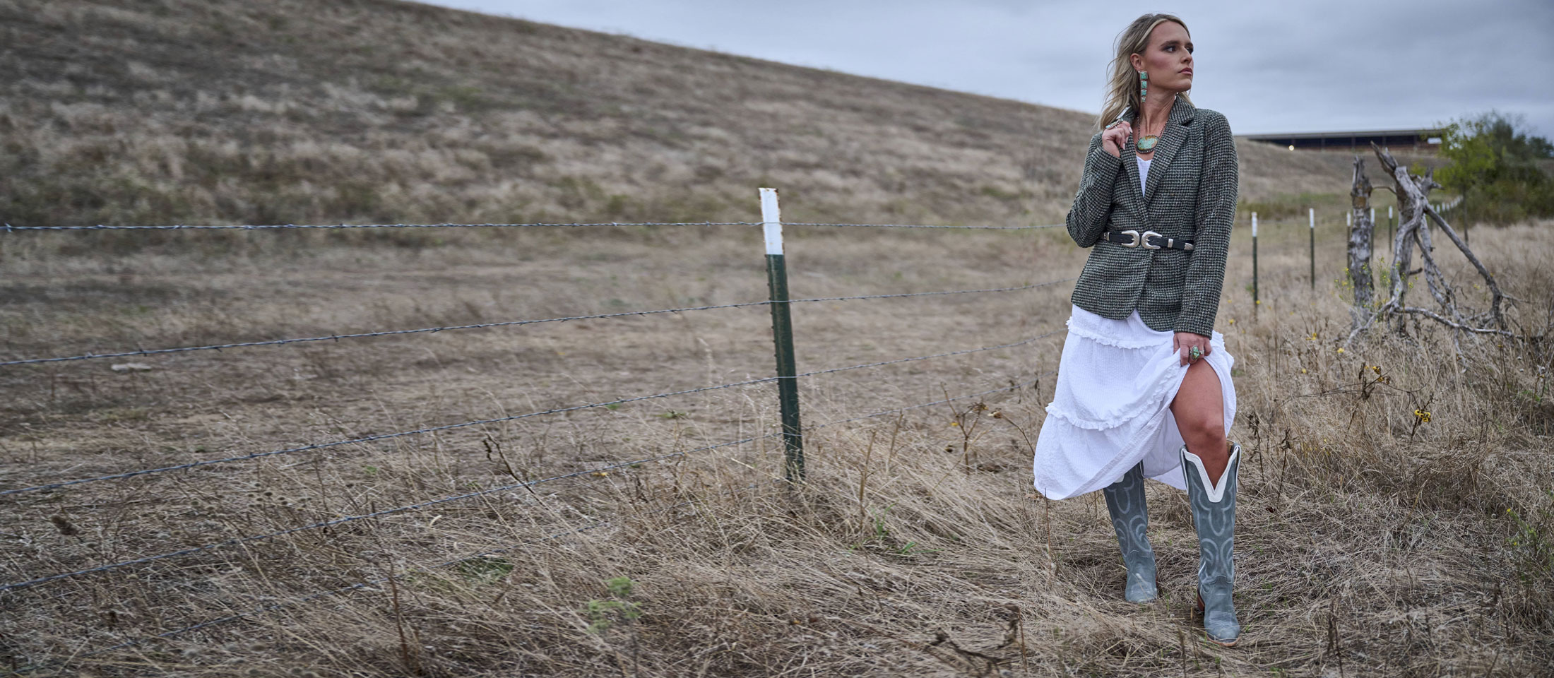 A woman standing in a pasture with tall grass, posing in a chic jacket, looking away from the camera.